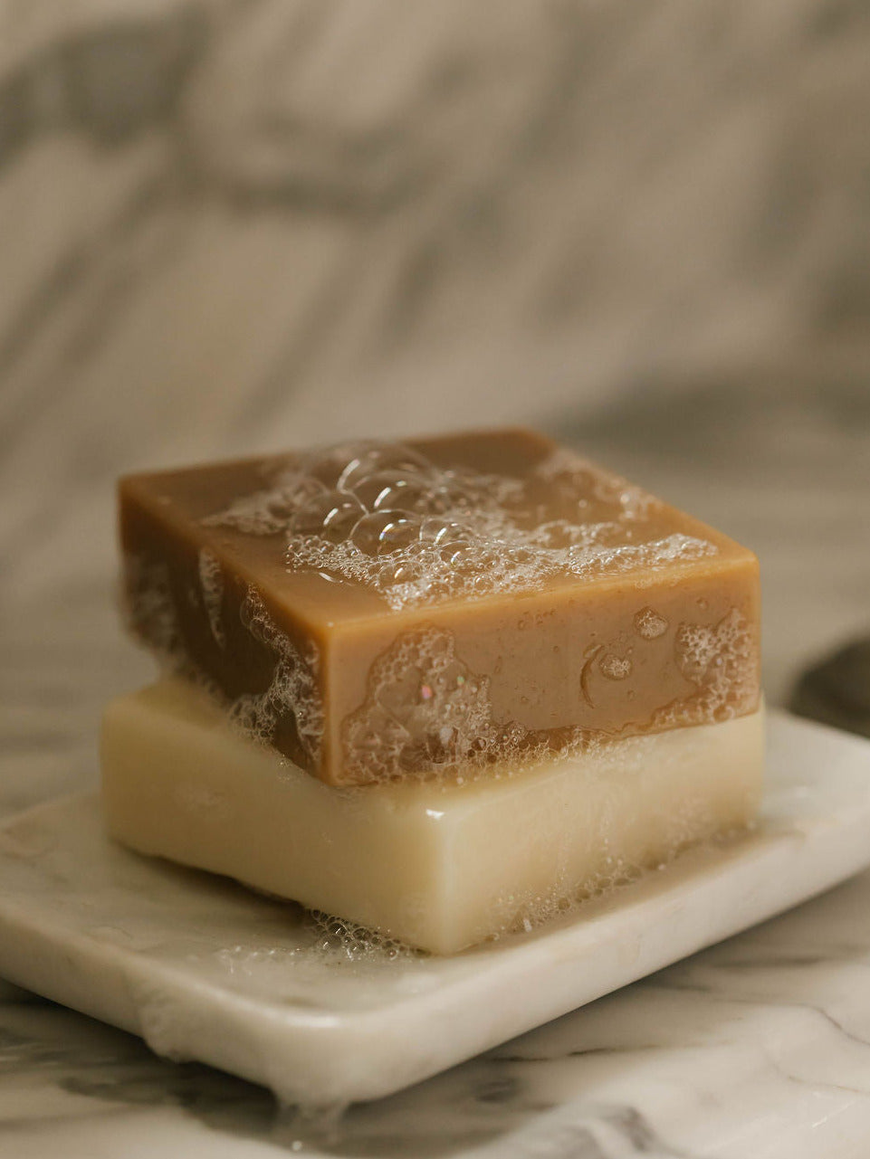 Own Skin's square bars of soap stacked on a small white marble dish, set against a softly blurred marble surface. The top bar is medium brown with bubbles and the bottom bar is pale cream.
