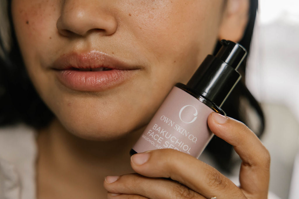 A close-up of a woman holding an Own Skin Co Bakuchiol Face Serum bottle. The bottle is a dusty mauve-pink color with a black pump dispenser, held against her cheek and jawline.