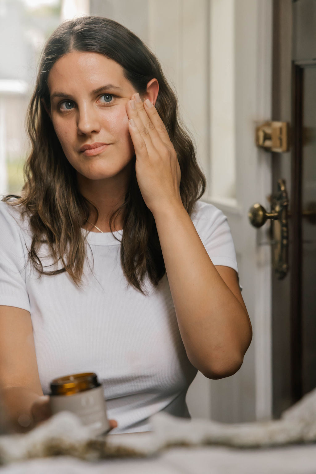 A woman with long brown hair applies beef tallow moisturizer from OWN SKIN CO in a small amber jar to her cheek in a bathroom setting.