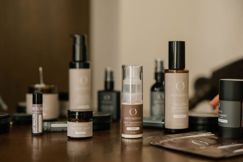 A collection of premium beef tallow skincare products from OWN SKIN CO. including dry shampoo, body oil, deodorant and lotions, arranged on a dark wooden table with a softly blurred indoor background.