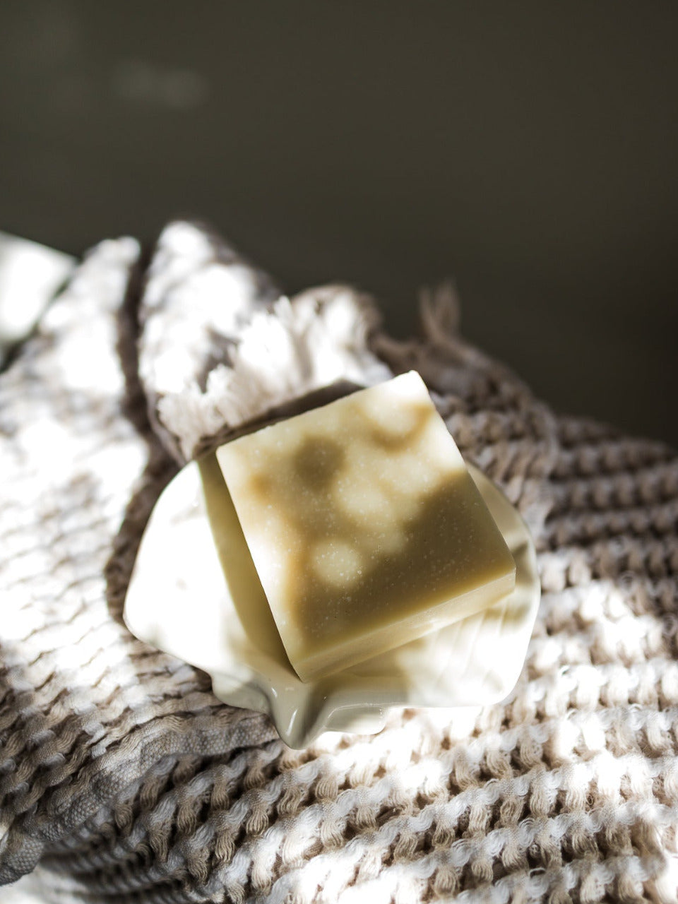 A cold process eucalyptus tallow square bar of soap, resting on a white ceramic shell shaped dish atop a textured, woven beige and white cloth under dramatic, dappled sunlight.