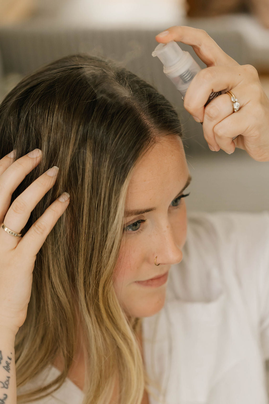 A woman with long blonde-highlighted hair sprays her hair with natural dry shampoo from OWN SKIN CO. a clear pump bottle. Her hands are in her hair, and she is wearing a white top