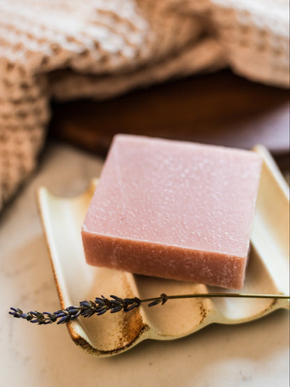 A square bar of dusty pink beef tallow body wash in a rose geranium floral scent resting on a small beige ceramic dish with a dried lavender sprig, set against a softly blurred background with a woven towel.