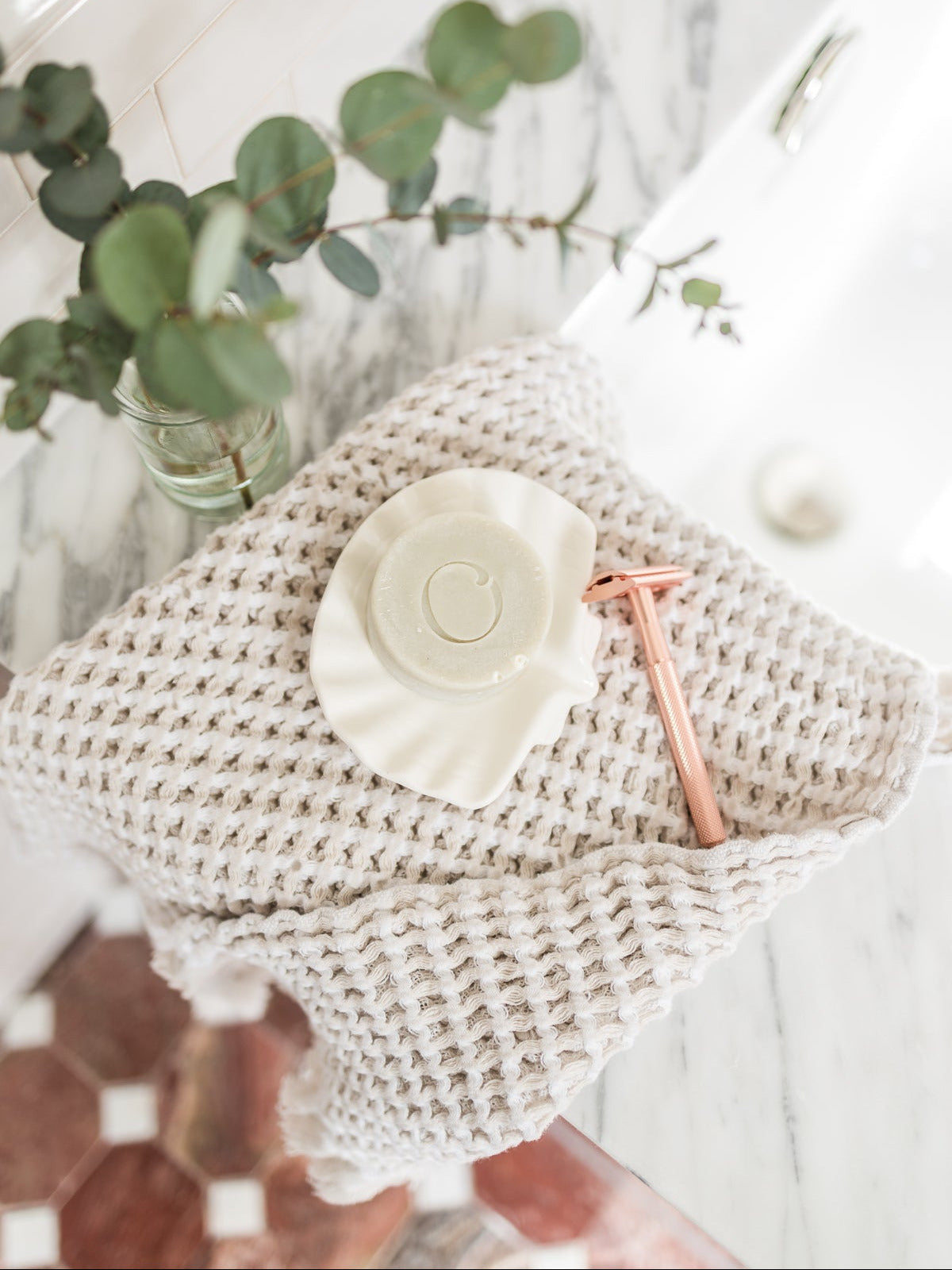 A close-up of a solid shave bar and a shaver resting on a waffled tallow on the side of a bathtub with a few sprigs of eucalyptus and a red tile floor in the background. 