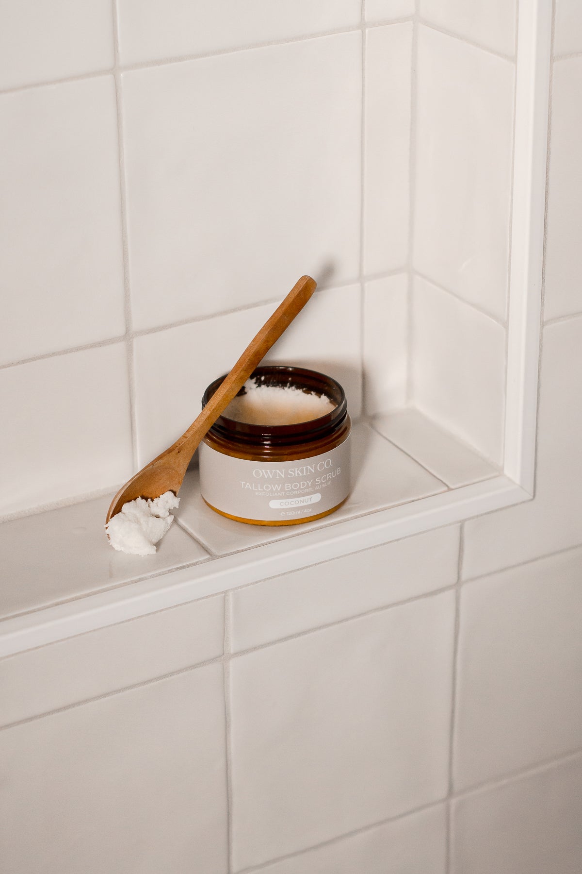 Jar of whipped beef tallow body scrub with a wooden spoon on a white shower ledge