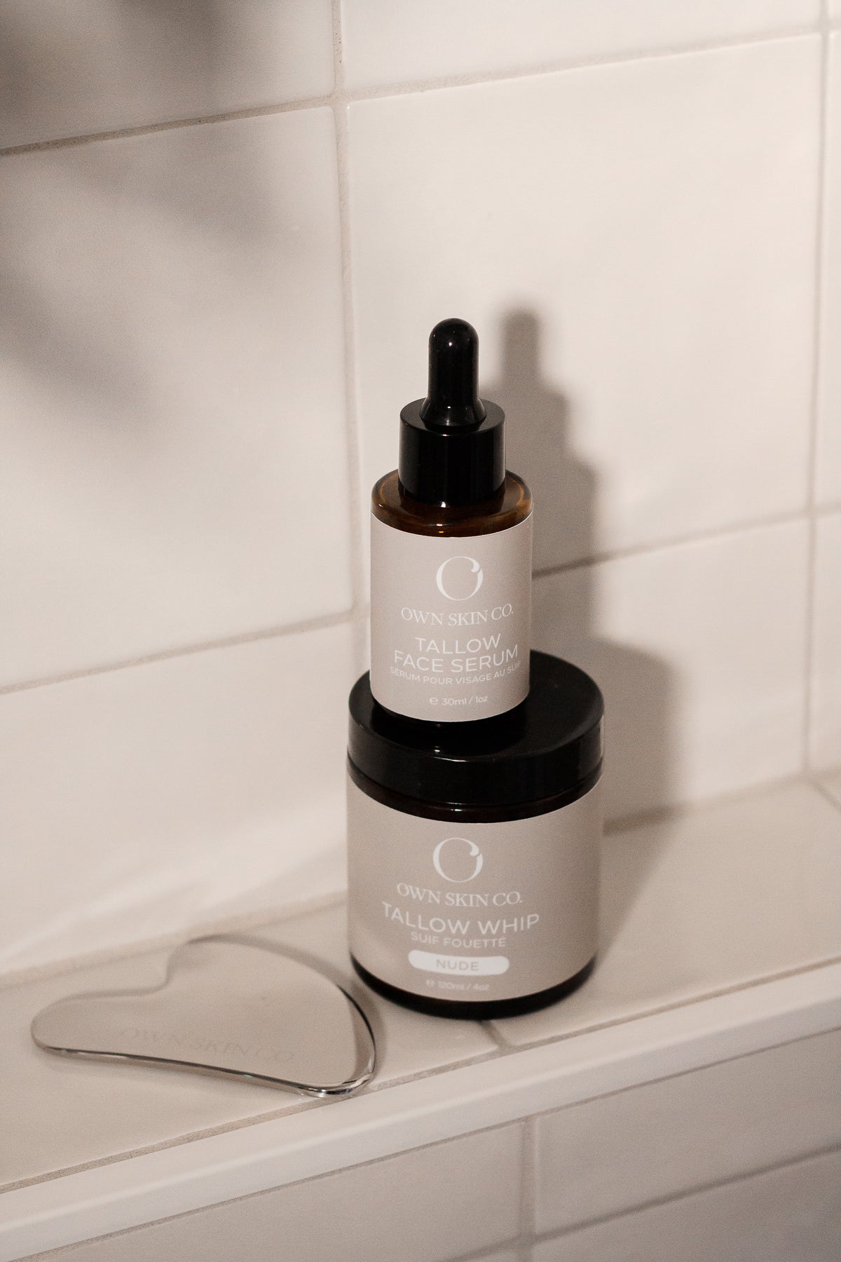 Beef Tallow face skincare tallow serum and whipped tallow moisturizer with own skin co stainless steel gua sha on shower ledge with white tiles