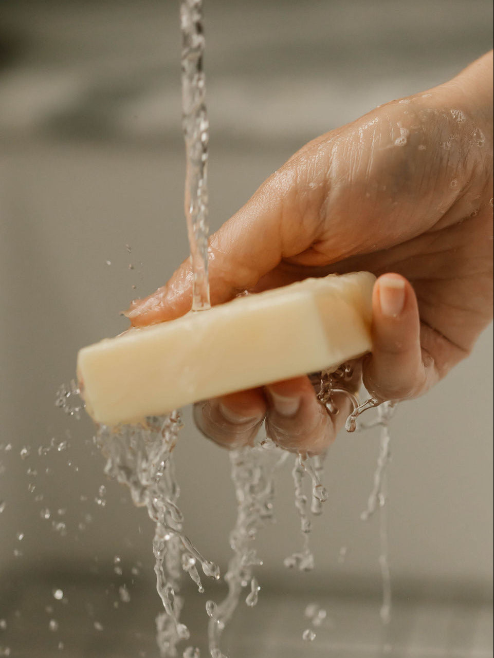 A hand holding a square bar of unscented solid body wash and hand soap from own skin co.'s soap line. It is under running water in a sink, with water droplets splashing around.