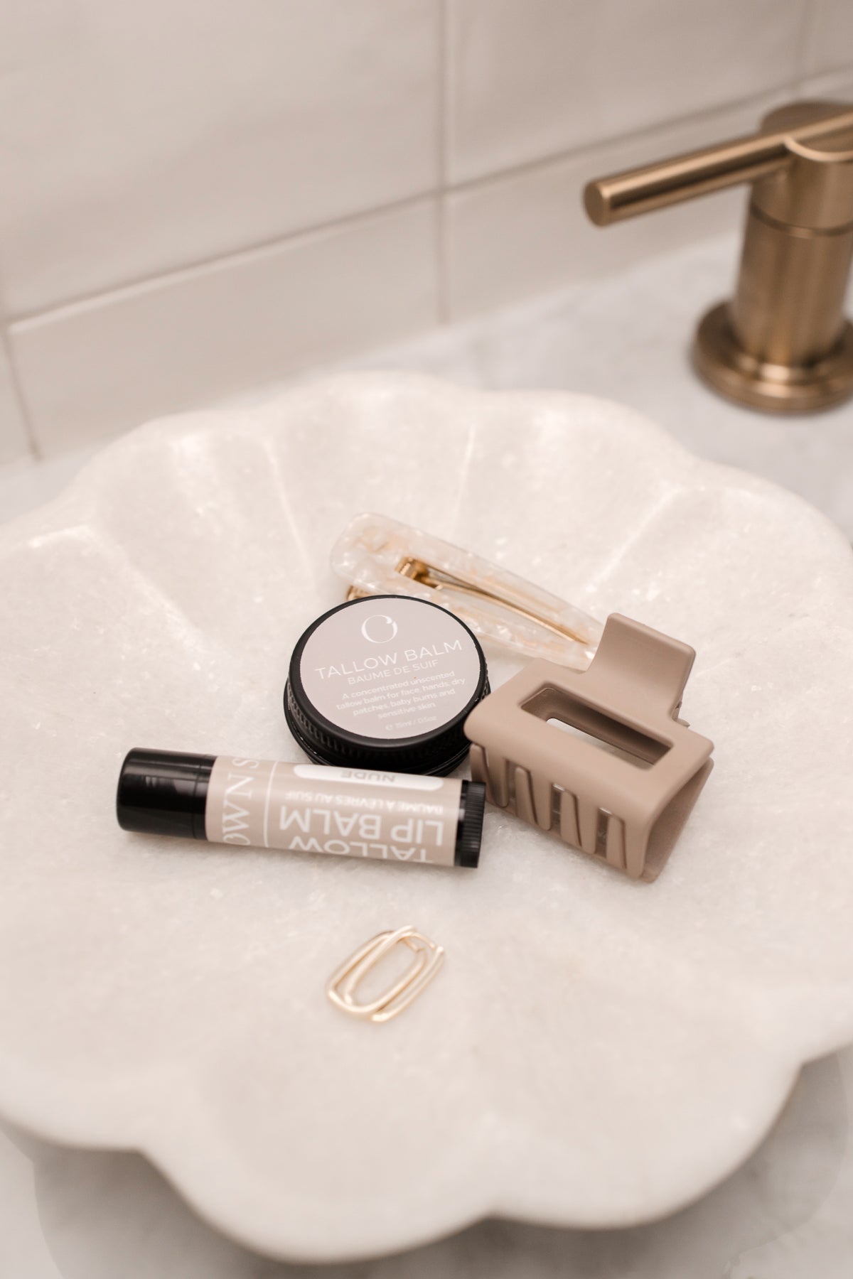 A white stone bowl with a natural unscented skincare balm, lip balm and gold earings and a beige hair clip