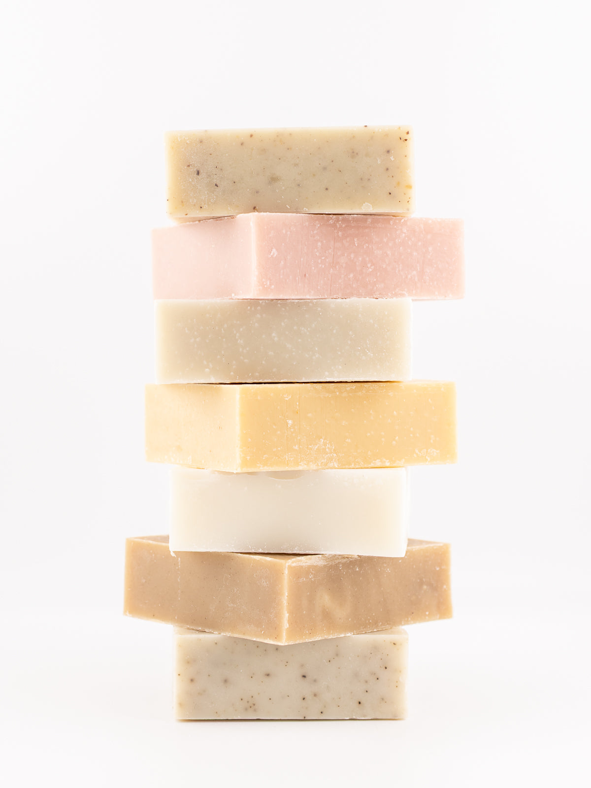 A vertical stack of seven square handmade beef tallow soap bars from OWN SKIN CO in assorted pastel and neutral shades, including beige, pink, cream, and light brown, photographed on a seamless white background.