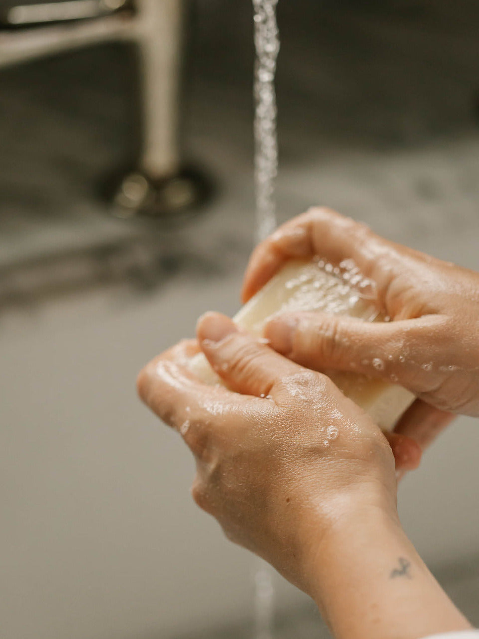 Close-up of hands washing with a bar of own skin co's soap under running water in a sink.