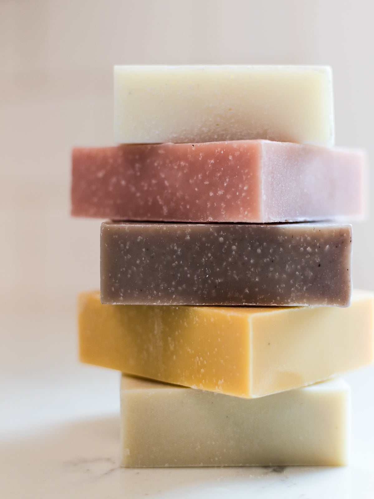 A stack of five cold process made in canada square solid body wash bars of soap in assorted earthy colors, including cream, dusty rose, taupe, mustard yellow, and off-white, photographed on a light neutral surface.