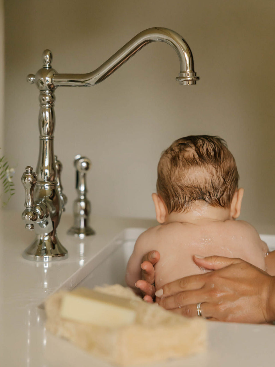 A baby sitting in a sink bathtub with an adult's hands supporting them, featuring a tall polished chrome faucet in the background and OWN SKIN CO natural baby soap bar in a dish on the counter. 