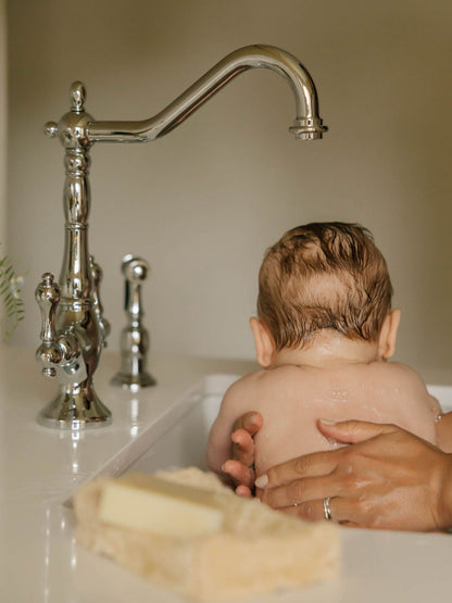 A baby sitting in a sink bathtub with an adult's hands supporting them, featuring a tall polished chrome faucet in the background and OWN SKIN CO natural baby soap bar in a dish on the counter. 