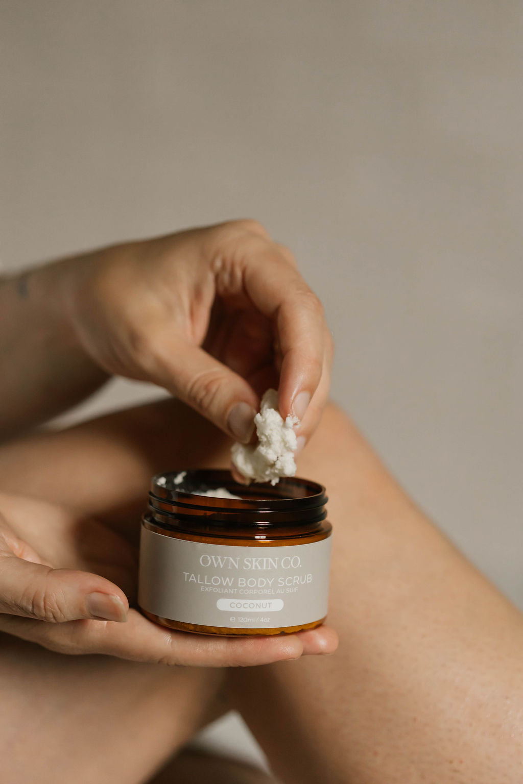 A person holds an amber glass jar of Own Skin Co. tallow Body Scrub while dipping their hand into the white scrub, with their legs visible in the background against a neutral beige wall.