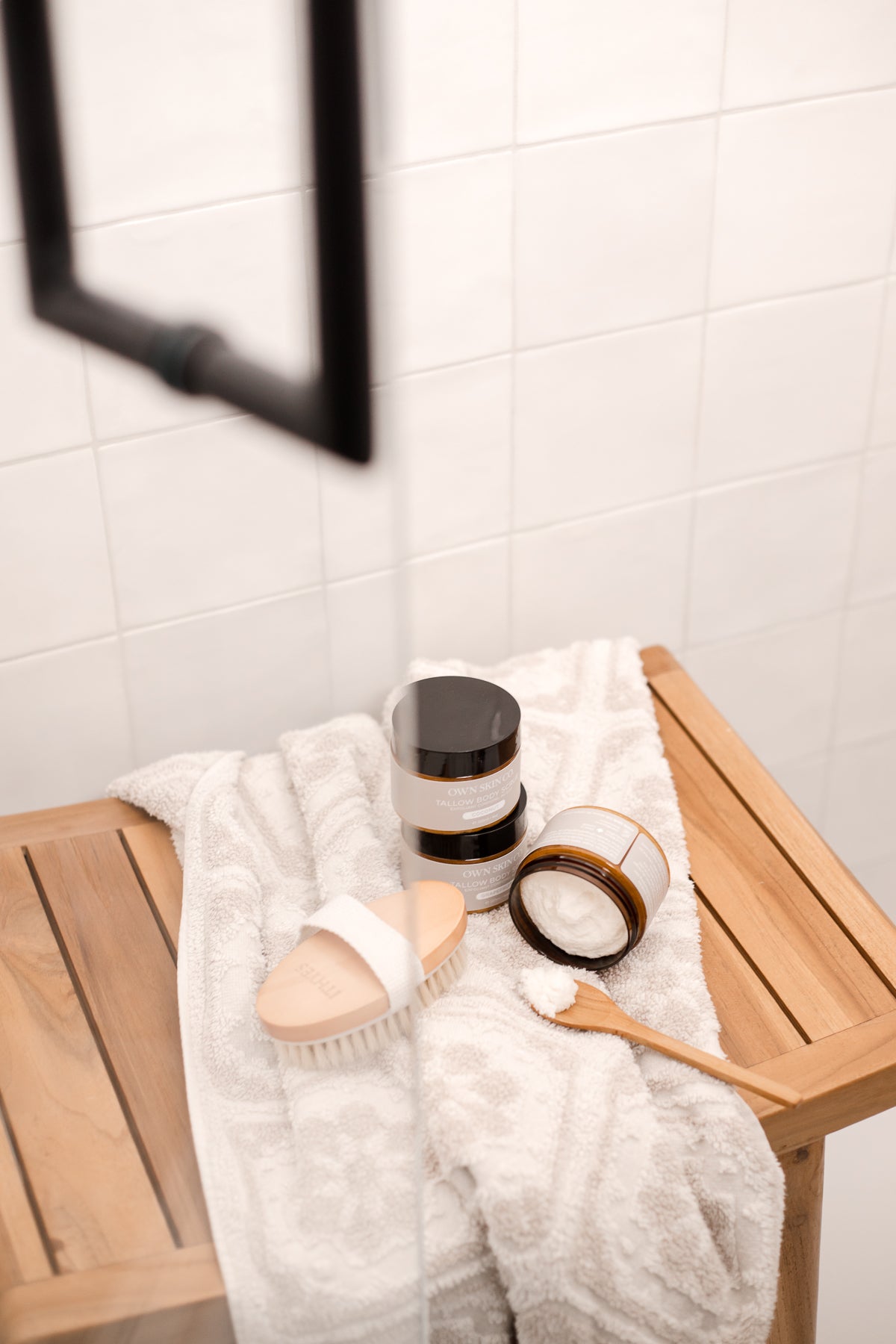Beef Tallow Body Scrub with open jar and spoon on a towel in a shower with a dry brush on a bench