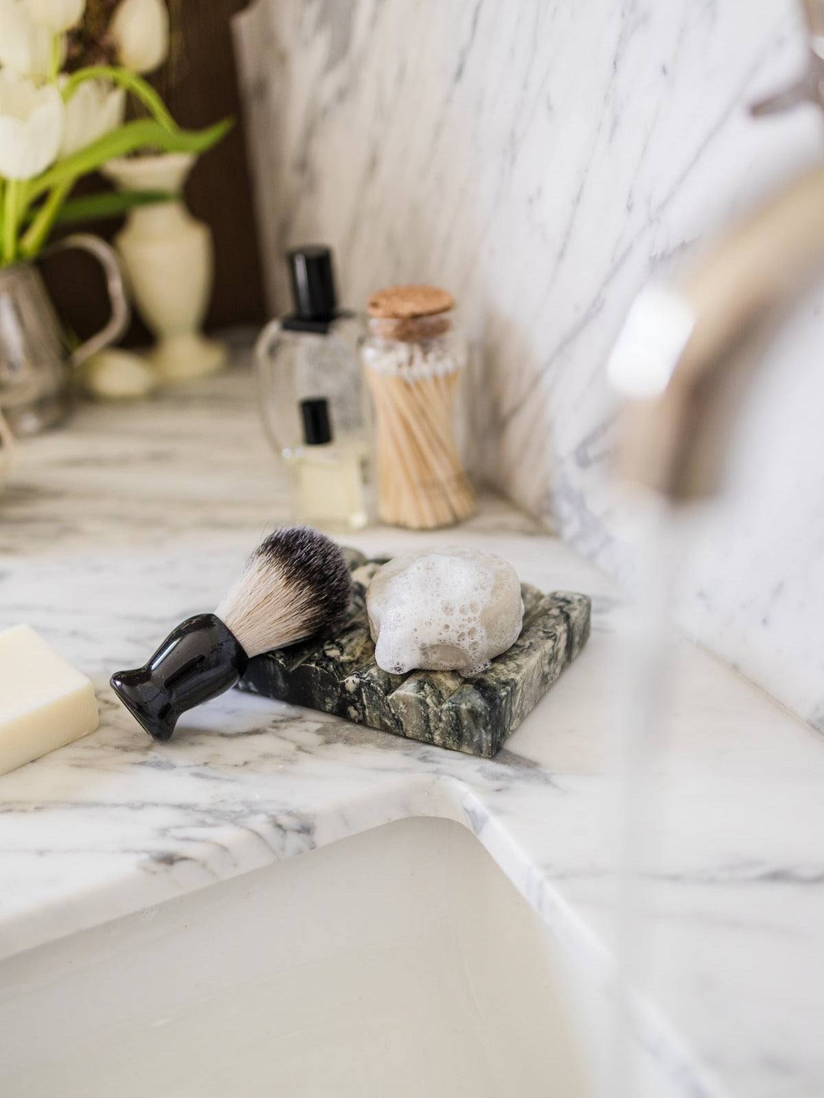 A shaving brush with black bristles and a black handle rests on a dark marble soap dish beside an own skin co men's tallow shave bar