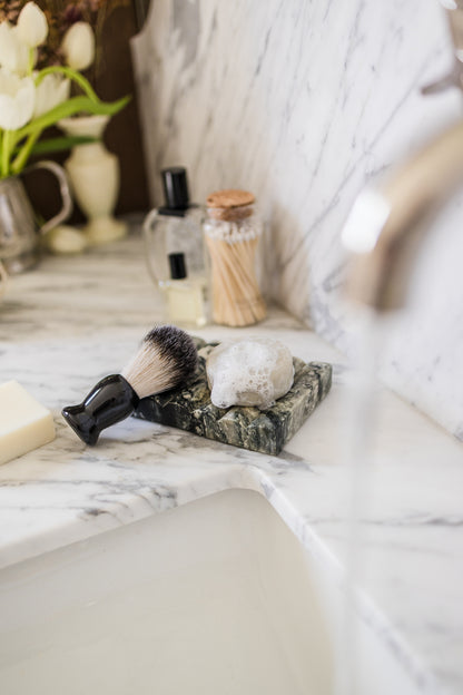 A shaving brush with black bristles and a black handle rests on a dark marble soap dish beside an OWN SKIN CO men's solid shaving bar with bubbles on it. Set on a white marble bathroom counter.