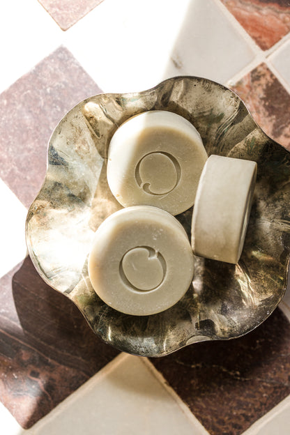 Three round solid shave bars made with beef tallow in canada in citrus scent. The shave bars are cream-colored an O engraved into them in the center, arranged in a scalloped metallic dish on a tiled surface.