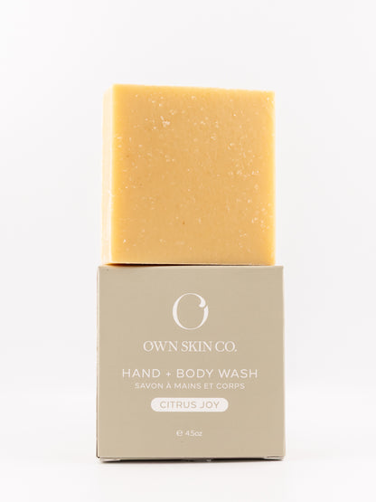 A rectangular bar of hand and body wash in a warm citrus yellow sits on top of its beige packaging box. The box features the brand name 'OWN SKIN CO.