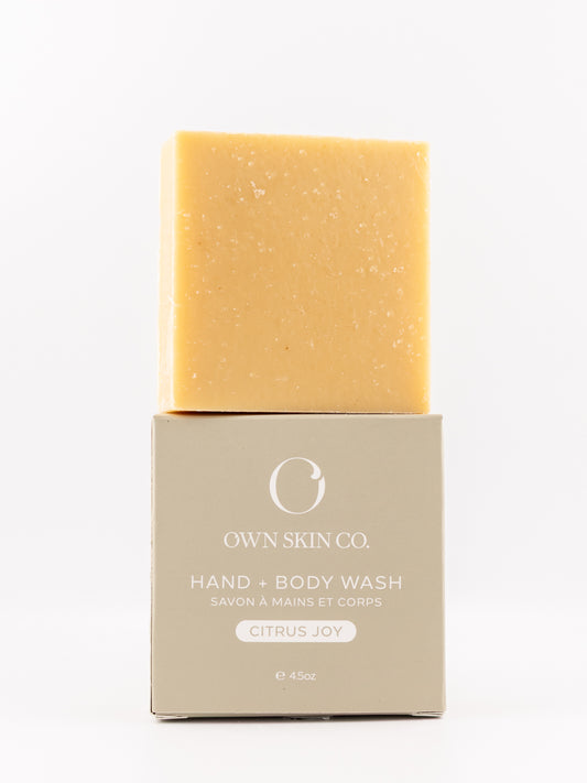 A rectangular bar of hand and body wash in a warm citrus yellow sits on top of its beige packaging box. The box features the brand name 'OWN SKIN CO.