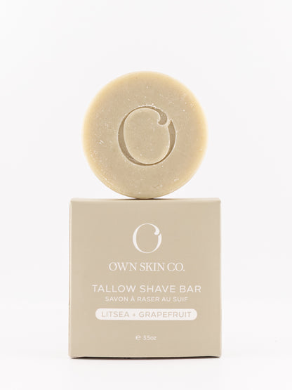 A round citrus tallow shave bar with an embossed 'O' logo sits on top of its beige packaging box. The box is labeled 'OWN SKIN CO.