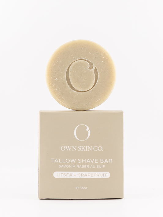 A round citrus tallow shave bar with an embossed 'O' logo sits on top of its beige packaging box. The box is labeled 'OWN SKIN CO.
