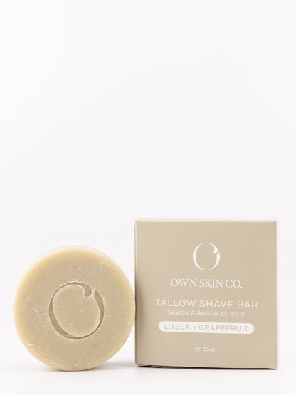 A round beige shave bar with an embossed logo sits next to its beige packaging box on a clean white background. The box is labeled 'OWN SKIN CO.' litsea and grapefruit scent