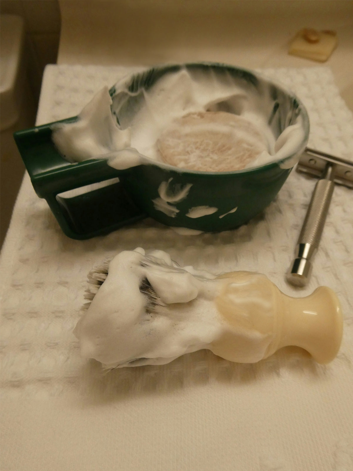 An own skin co men's solid shave bar in a dish that's been lathered into a rich cream with a shaving brush which is laying in front on a towel, covered with shaving foam from the bar.