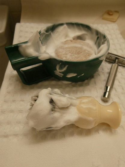 An own skin co men's solid shave bar in a dish that's been lathered into a rich cream with a shaving brush which is laying in front on a towel, covered with shaving foam from the bar.