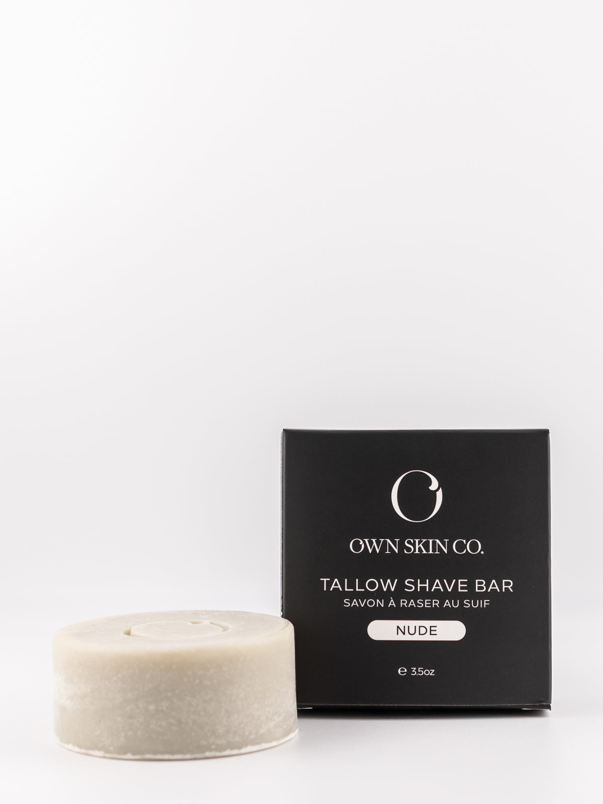 A round, textured off-white tallow shave bar sits next to its black packaging box, which features the 'Own Skin Co.' logo and 'Tallow Shave Bar' text, on a clean white background.
