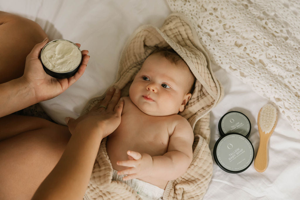 A baby wrapped in a beige textured blanket lies on a white bed while mom holds an open jar of tallow baby cream. Two OWN SKIN CO tallow baby balm jars and a wooden brush sit nearby on the bedding.