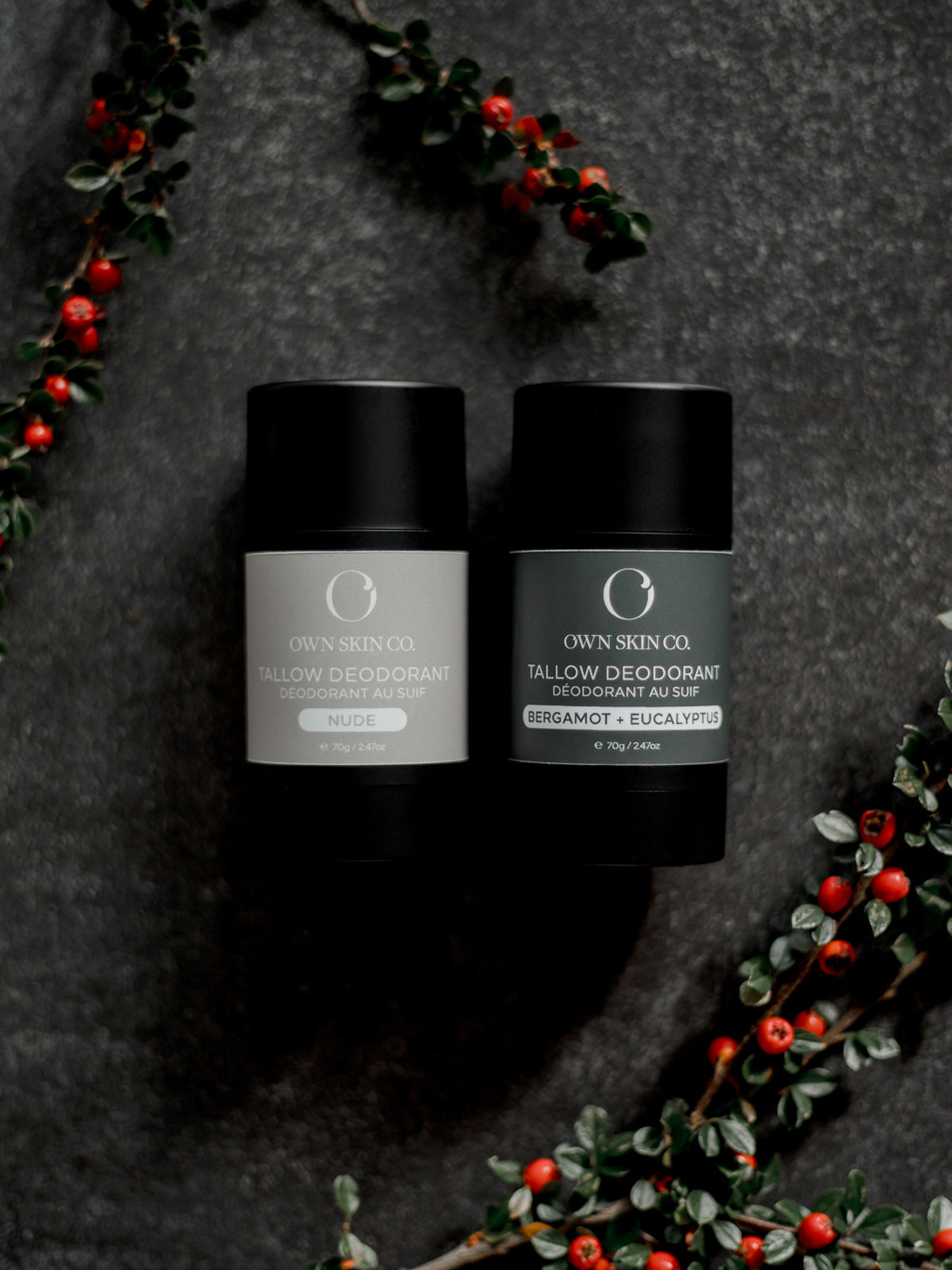 Two natural, tallow deodorant containers on a dark surface with decorative christmas greens and berries.
