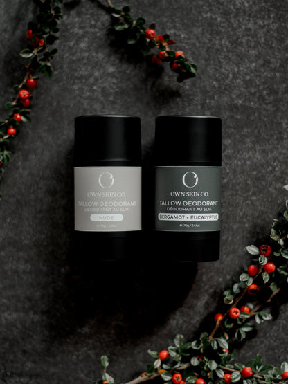 Two natural, tallow deodorant containers on a dark surface with decorative christmas greens and berries.