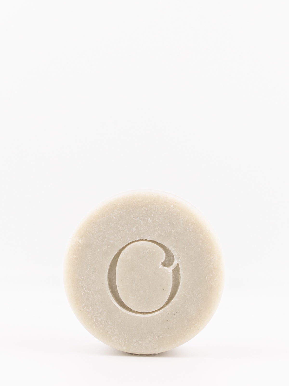 A single round bar of OWN SKIN's shaving soap with a carved letter 'O' in the center, shown on a seamless white background.