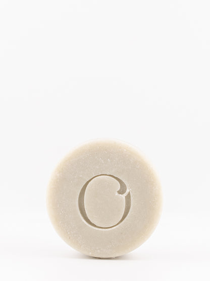 A single round bar of OWN SKIN's shaving soap with a carved letter 'O' in the center, shown on a seamless white background.